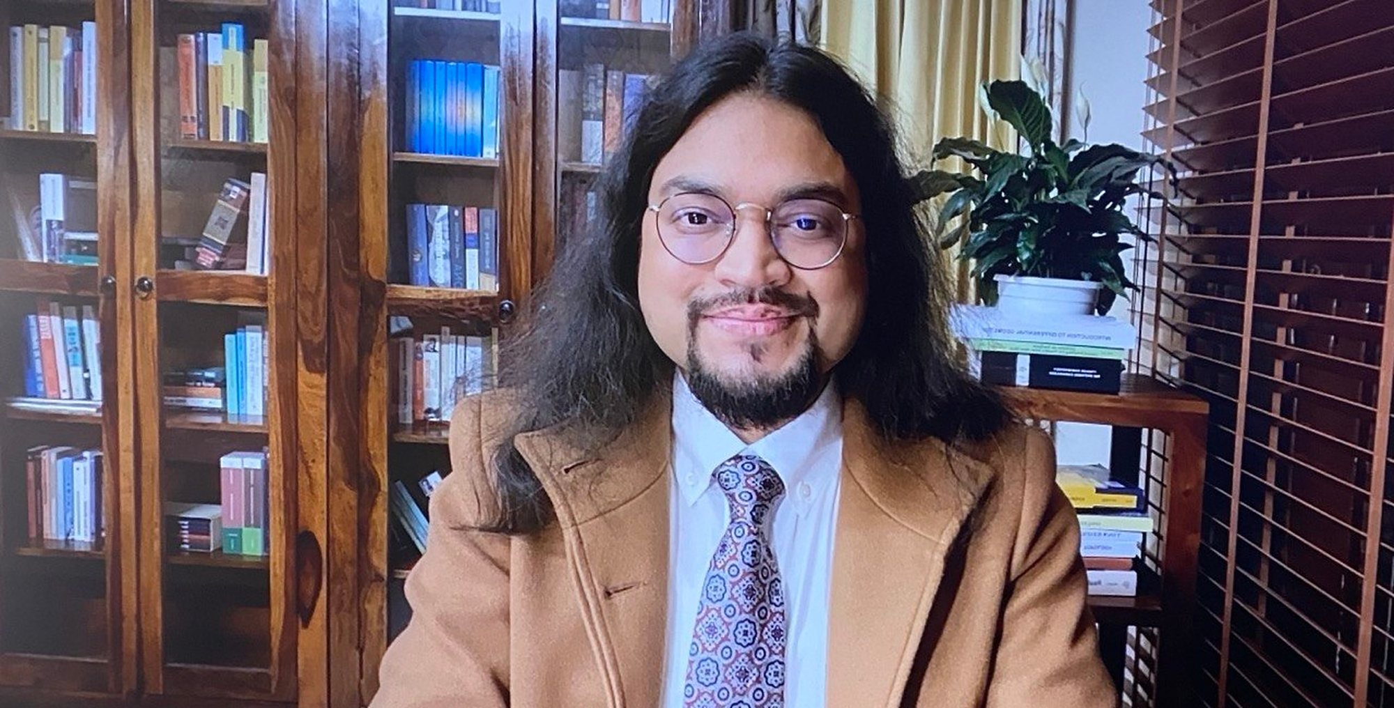 Yaashaa Golovanov in his study, in a camel coat, white shirt and patterned tie, before glass-fronted bookcases; a potted plant on a stack of books and wooden blinds to the right.